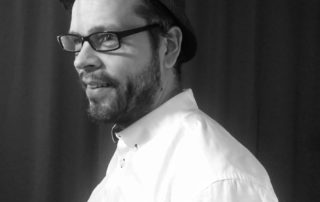 Berättaren Robert Markström iklädd skjorta, glasögon och hatt. Han ler och tittar år sidan.